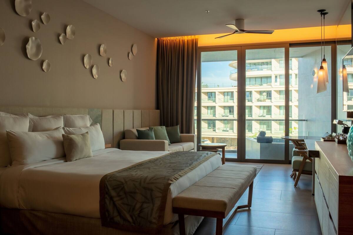 A hotel room with a large bed, a sofa, a bench, a desk, and a view of a building through a large window.