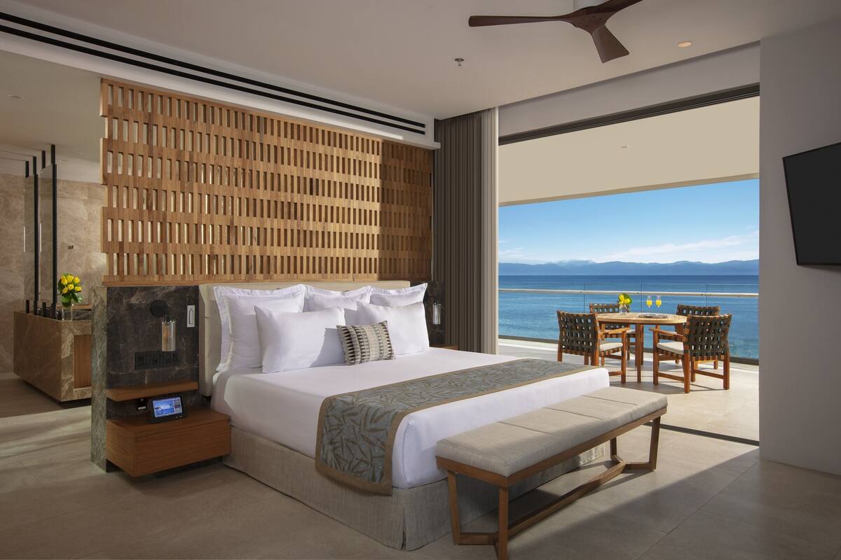 A modern hotel room with a large bed, a wooden headboard, a bedside table with a digital clock, a bench, and a balcony with a view of the sea.