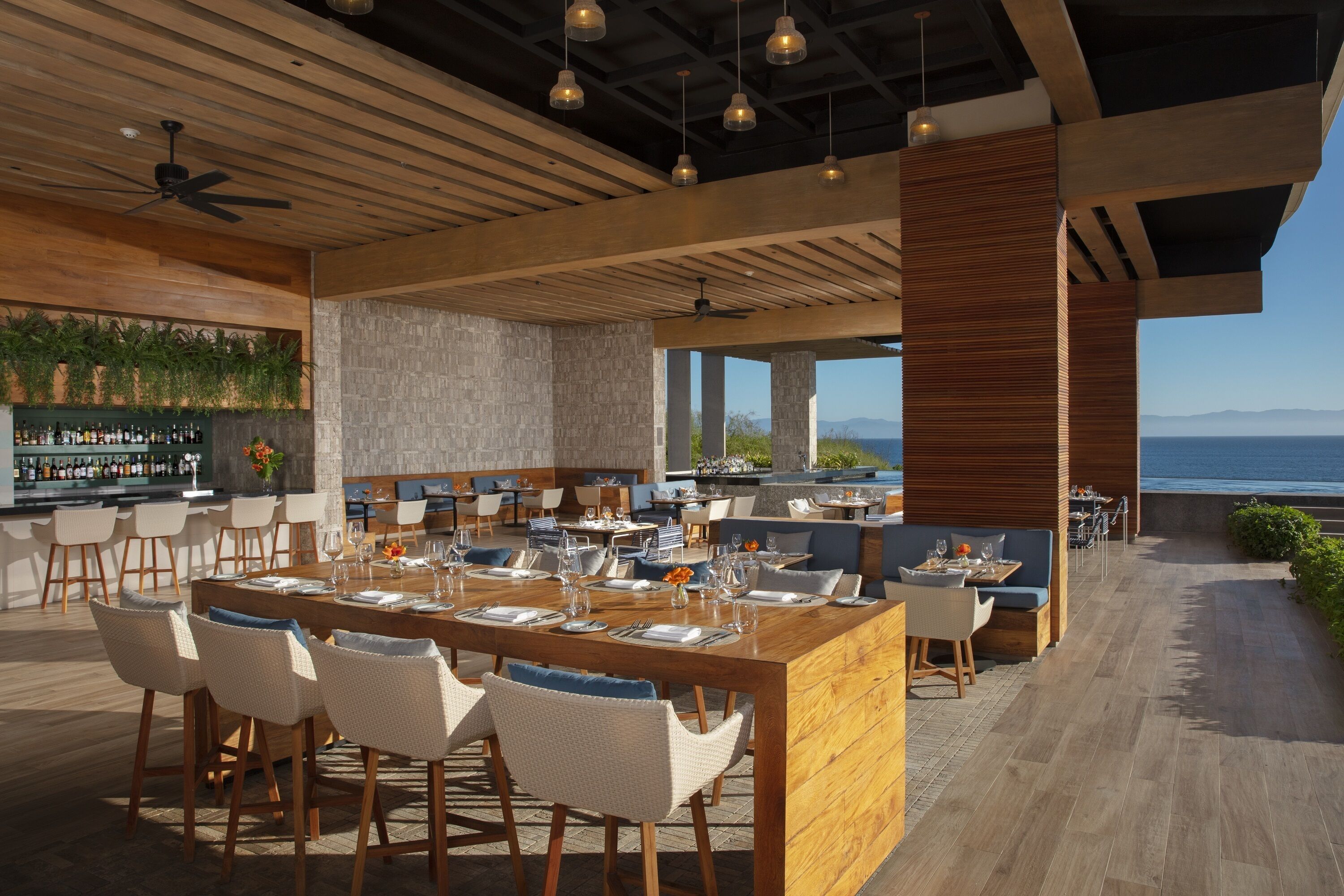 Un espacio moderno para comer al aire libre con barra, techo de madera y vistas al mar.