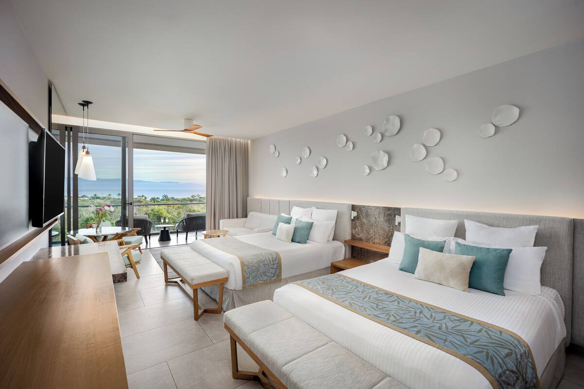 A modern hotel room with two beds, a dining area, and a large window with a view.
