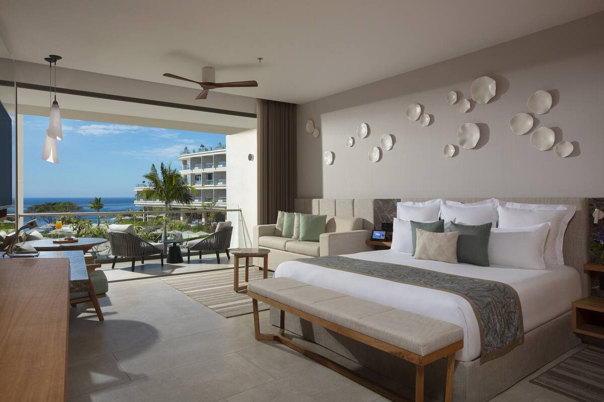 A modern hotel room with a large bed, a balcony with seating, and a view of the ocean.