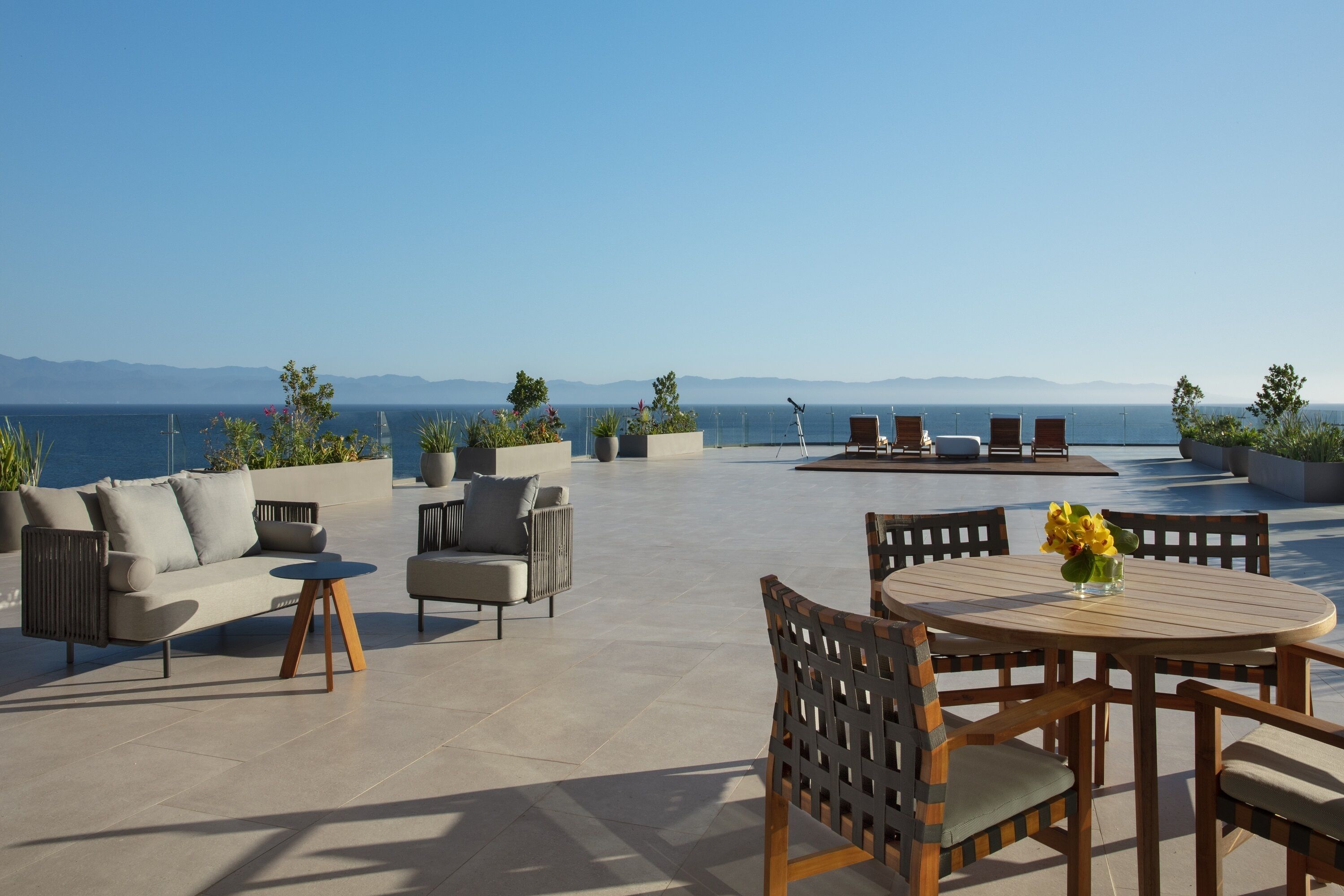 An outdoor patio with a round table and chairs, a sofa, and a lounge area with a view of the sea and mountains.