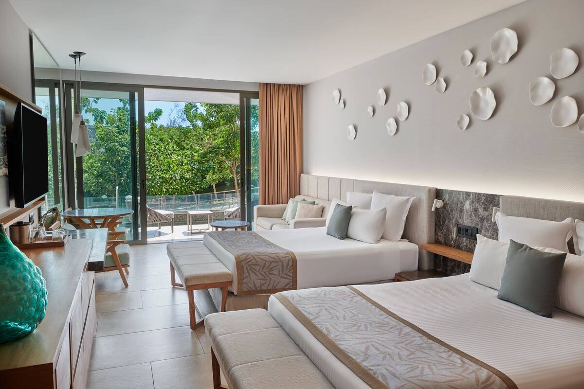 A hotel room with two beds, a TV, a dining area, and a balcony with a view of greenery.