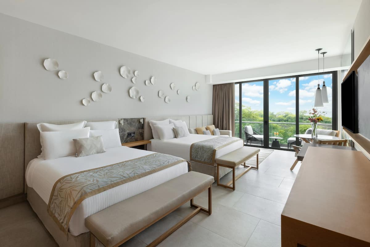 A hotel room with two beds, a desk, and a large window offering a view of greenery.