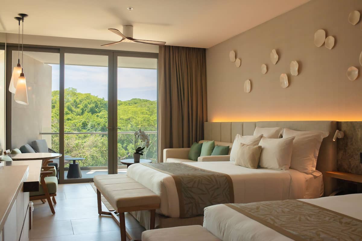 A modern hotel room with a large bed, a desk, and a balcony with a view of greenery.