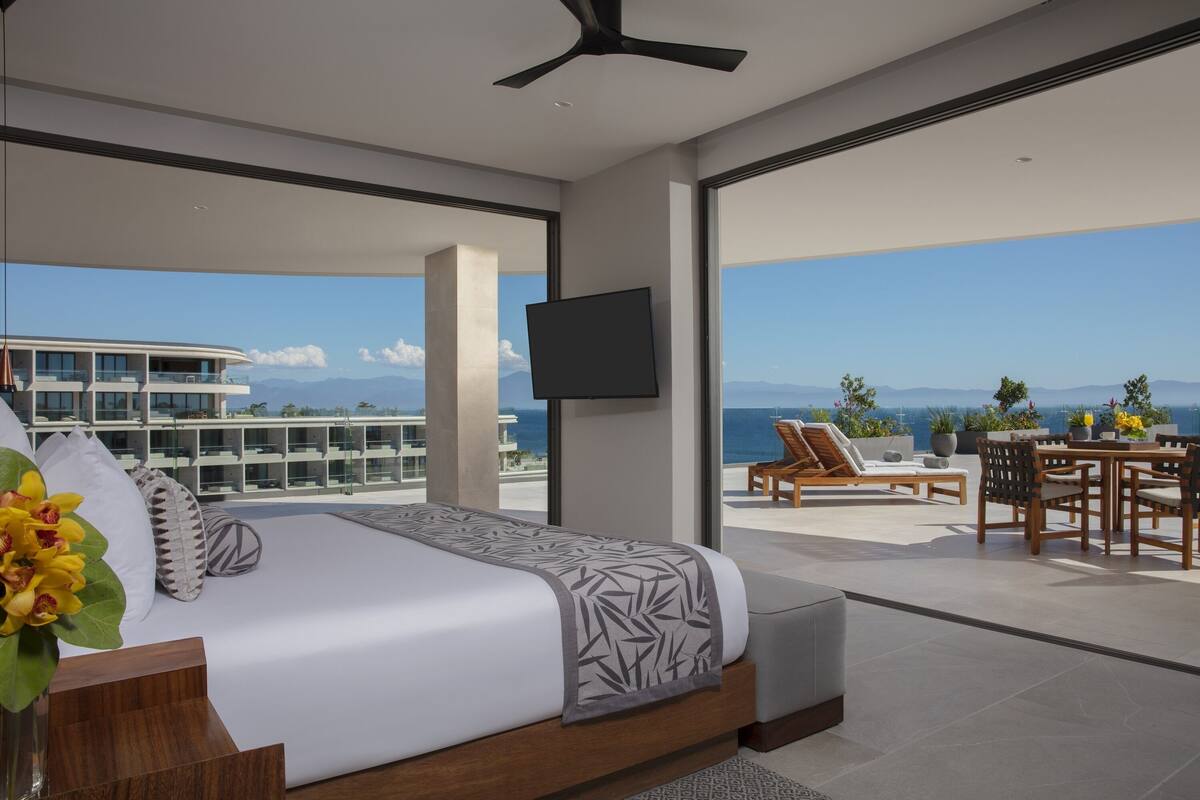 A modern bedroom with a large bed, a flat-screen TV, and a balcony with a view of the ocean and mountains.