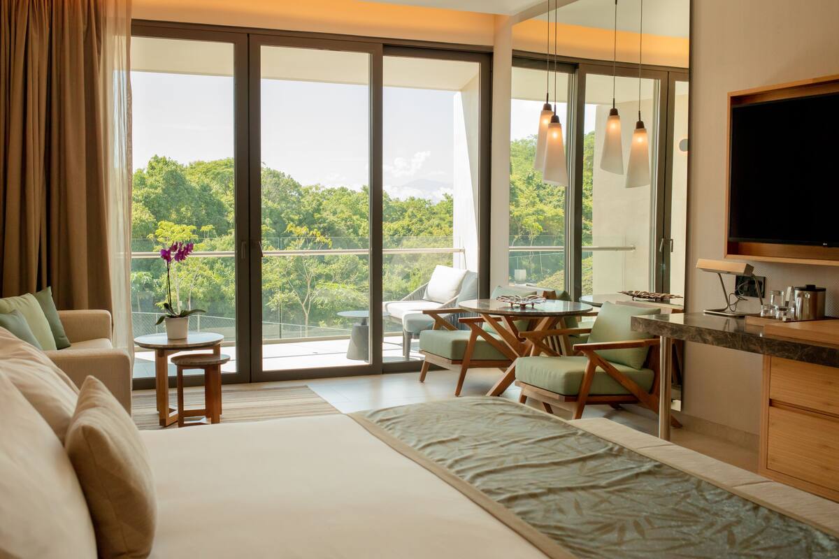 A modern hotel room with a large bed, a dining area, a TV, and a balcony with a view of the greenery.