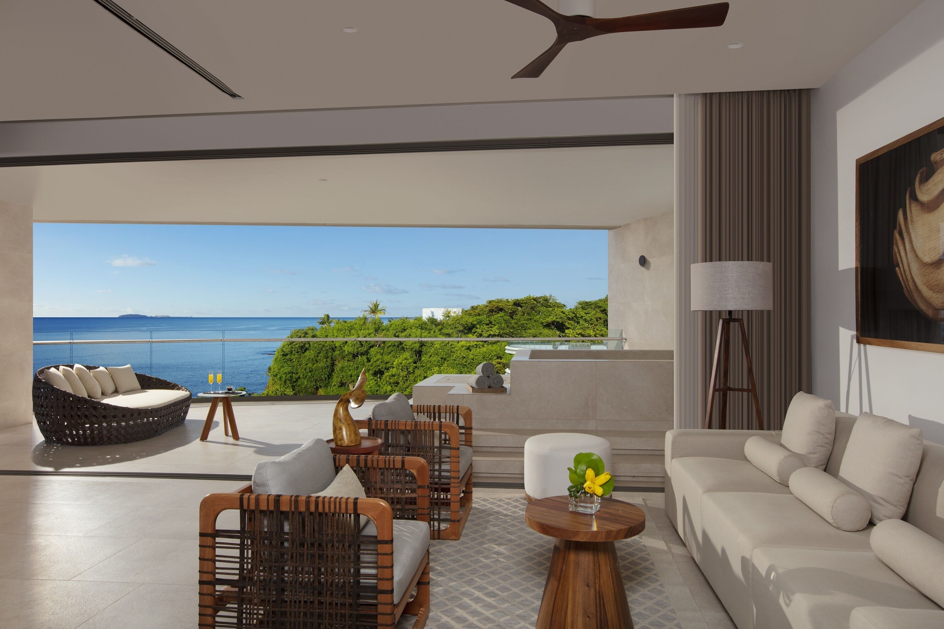 A modern living room with a sofa, chairs, and a coffee table. There is a view of the ocean and greenery outside.