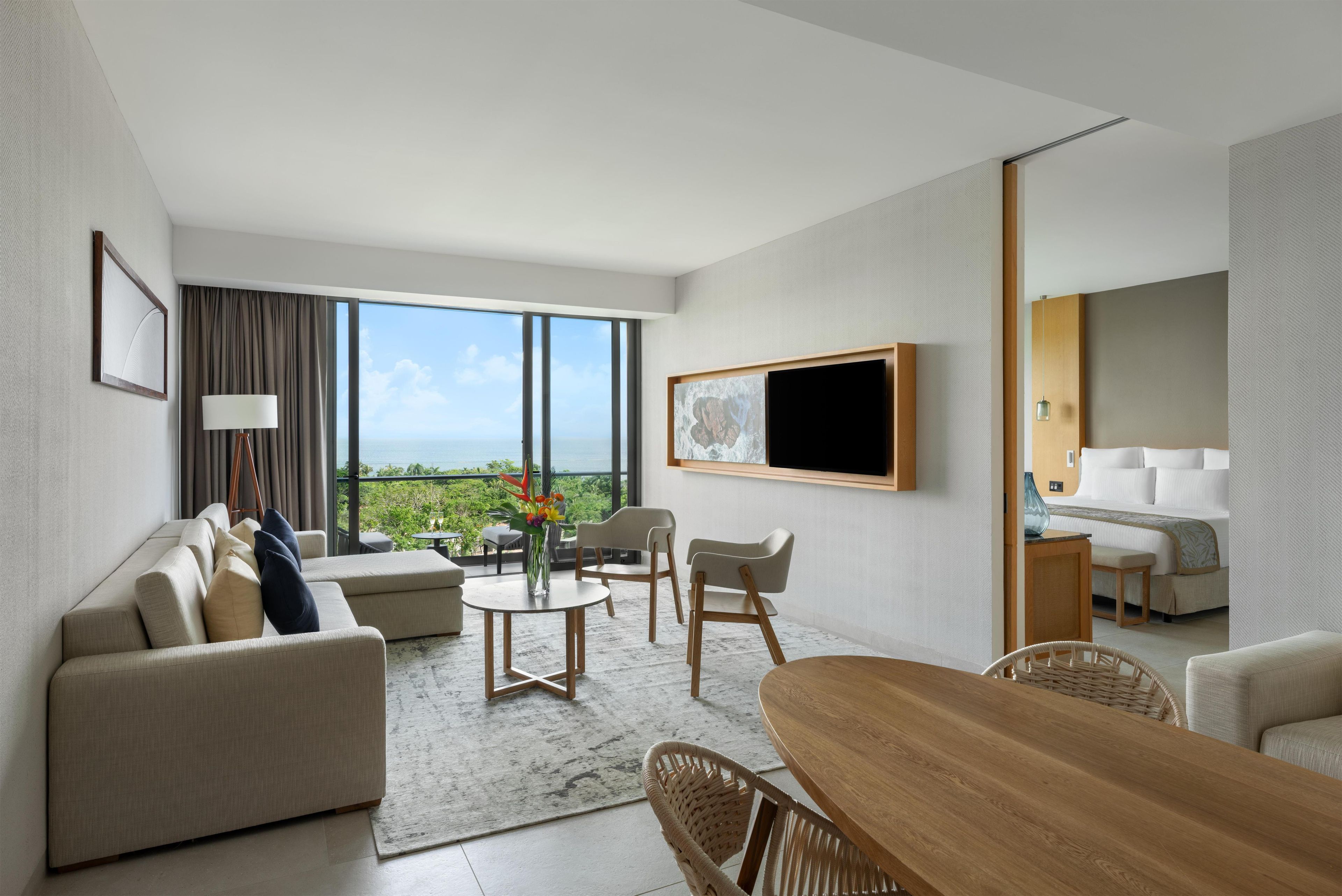 A modern hotel room with a large window, a flat-screen TV, a sofa, a dining table, and a bed.