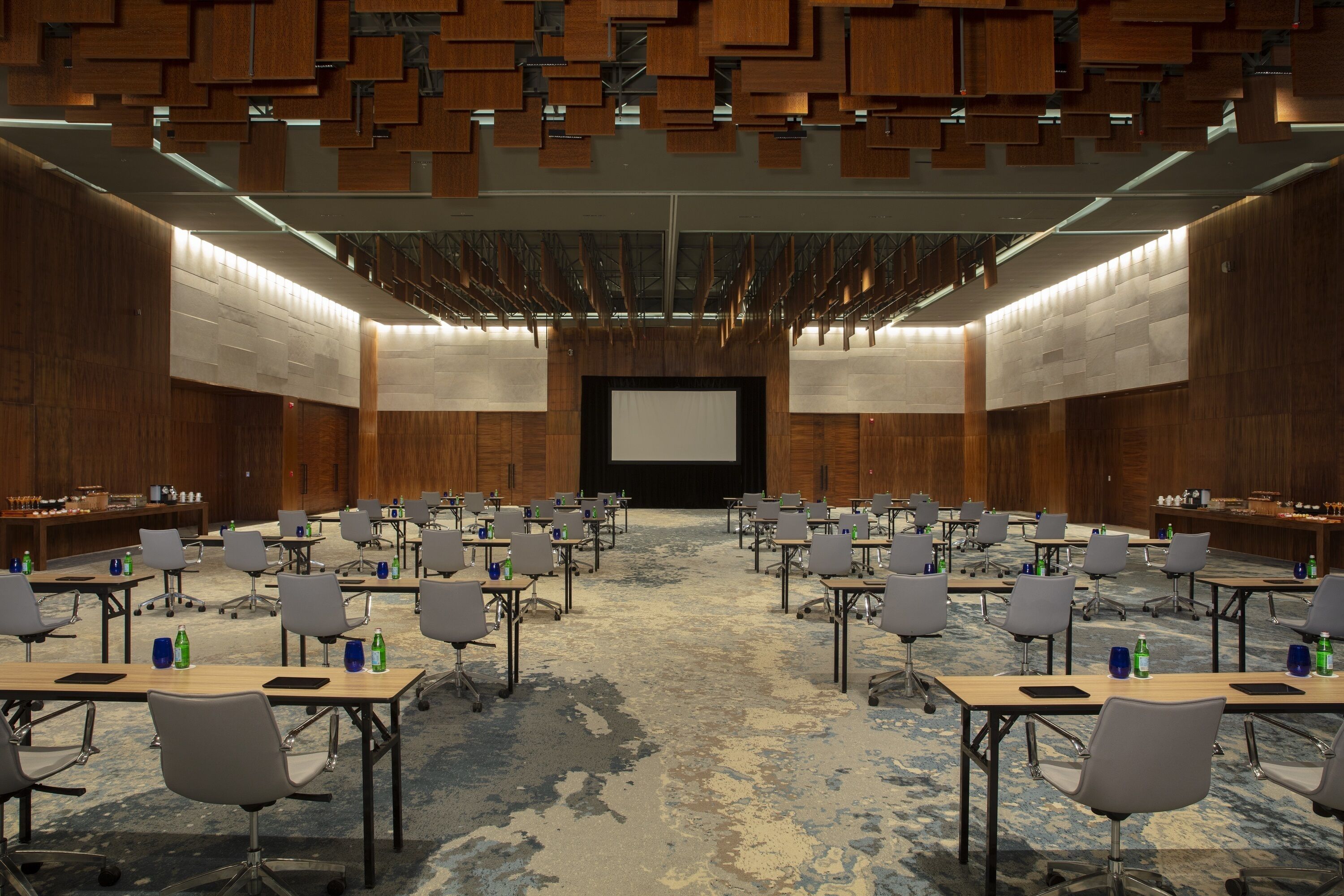 Une salle de conférence avec des rangées de tables et de chaises, un grand écran à l’avant et des panneaux de bois aux murs.