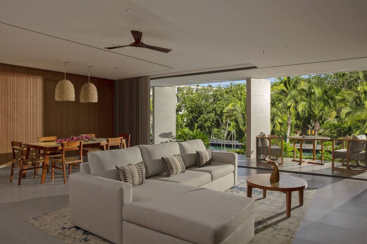 A modern living room with a sofa, dining table, and chairs. There is a ceiling fan and a view of lush greenery outside.