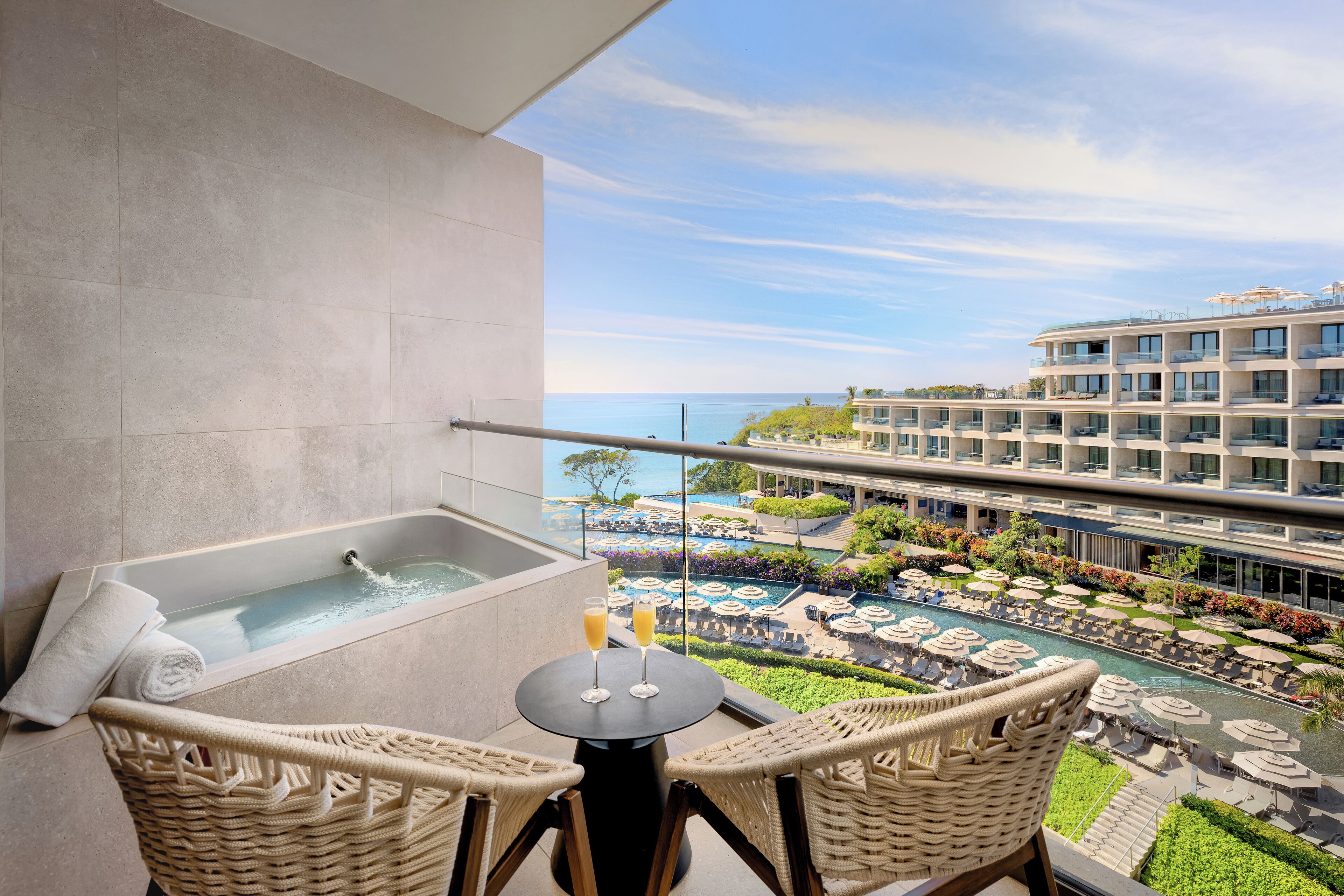A balcony with a bathtub, wicker chairs, and a view of a resort pool area.