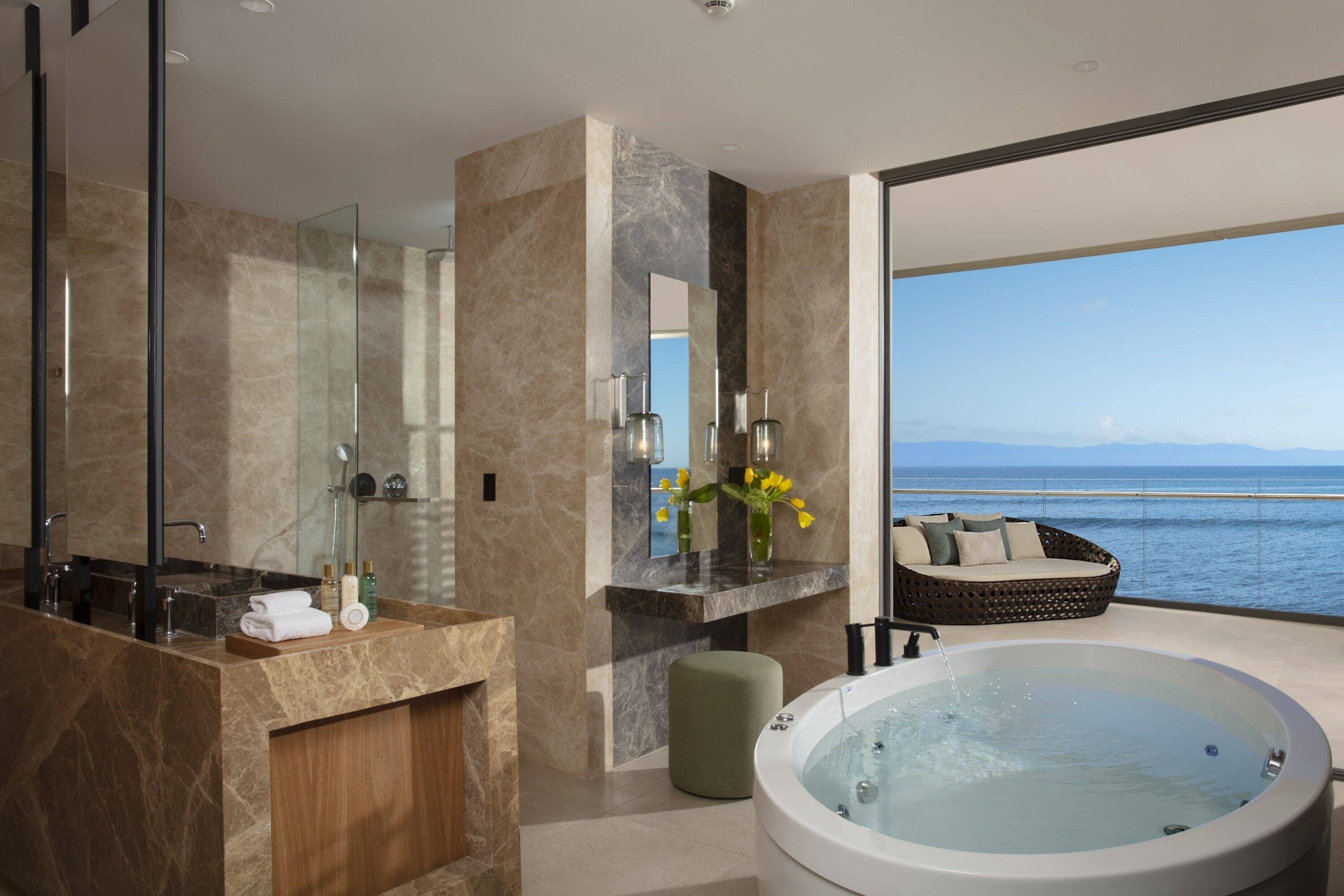 Un baño moderno con una bañera grande, cabina de ducha con paredes de vidrio y vista al mar.