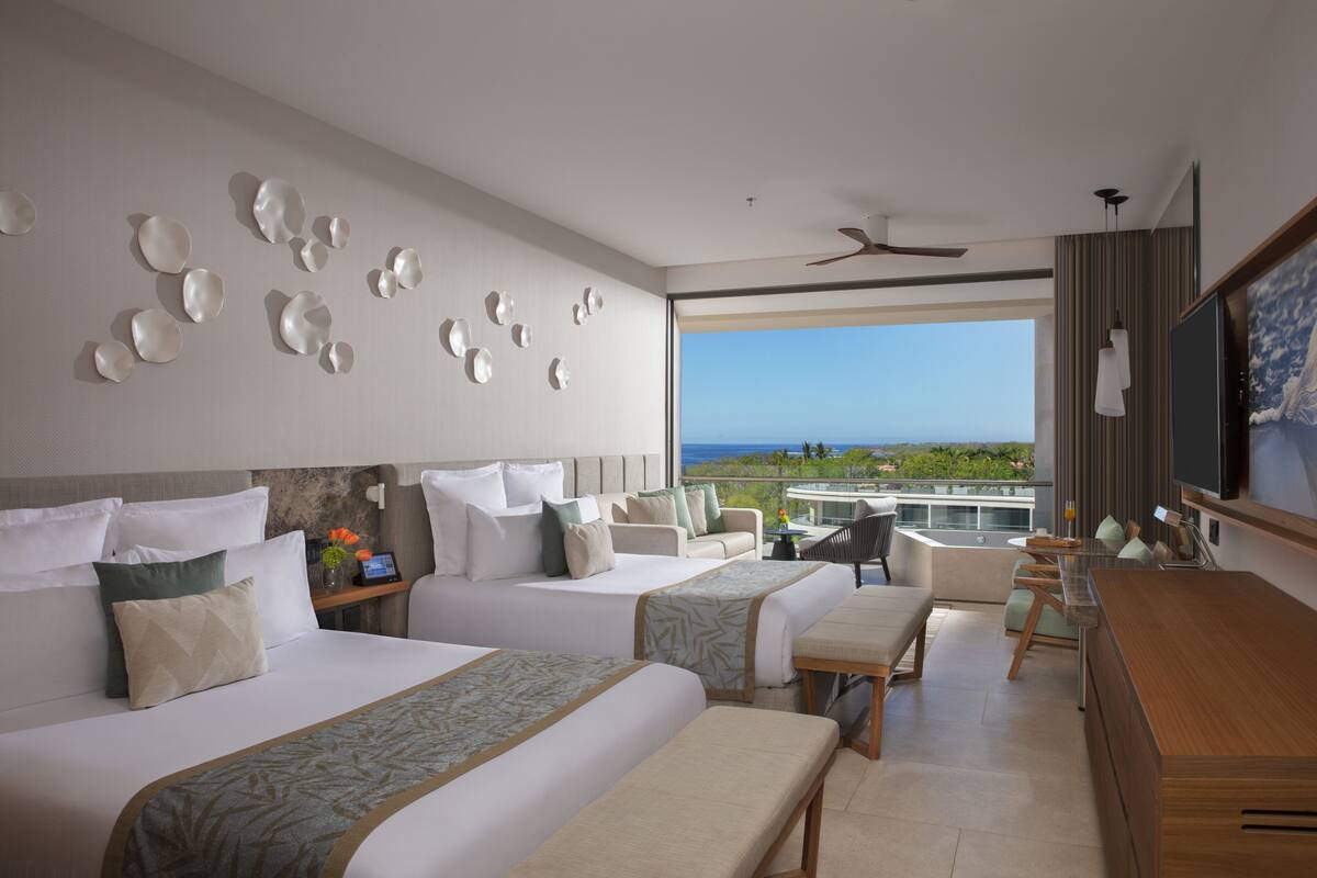 A hotel room with two beds, a desk, a chair, and a large window offering a view of the ocean.