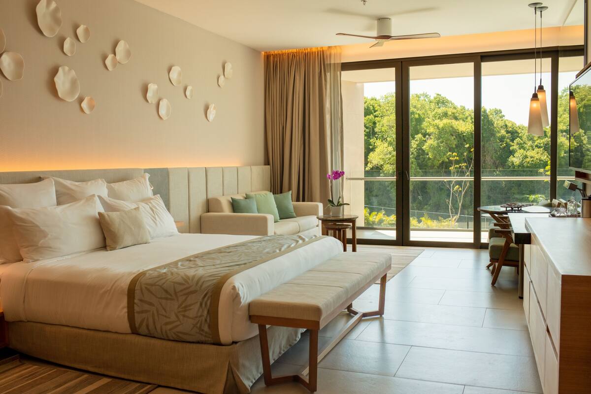 A modern hotel room with a large bed, a sofa, a dining area, and a balcony with a view of greenery.