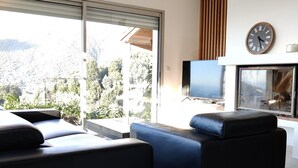A modern living room with a large sliding glass door, a flat-screen TV, a clock, and a fireplace. - Charming house with sea and mountain views and private pool (Sari-d'Orcino)