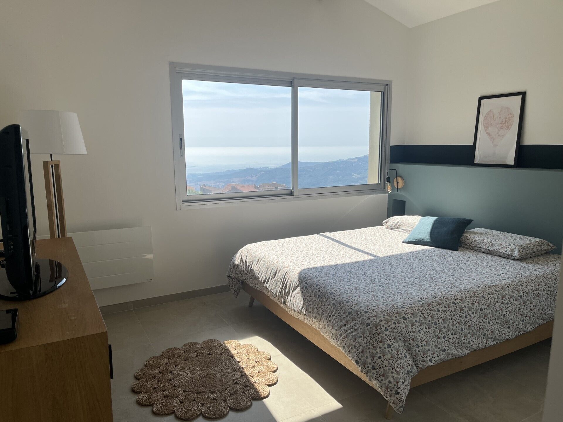 A bedroom with a bed, a desk, a lamp, a window with a view, and a framed picture on the wall.