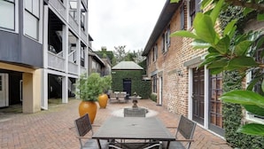 Une cour avec une table et des chaises, une fontaine et un bâtiment en briques dont la façade est également en briques, ainsi qu’un petit jardin.