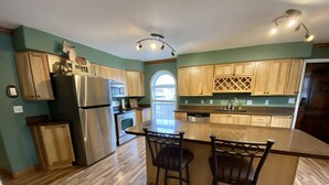 A kitchen with wooden cabinets, stainless steel appliances, a central island with bar stools, and a view through an arched window. - Western Carriage House Studio Suite- Western themed retreat- Full Kitchen & Bath (Gillette)