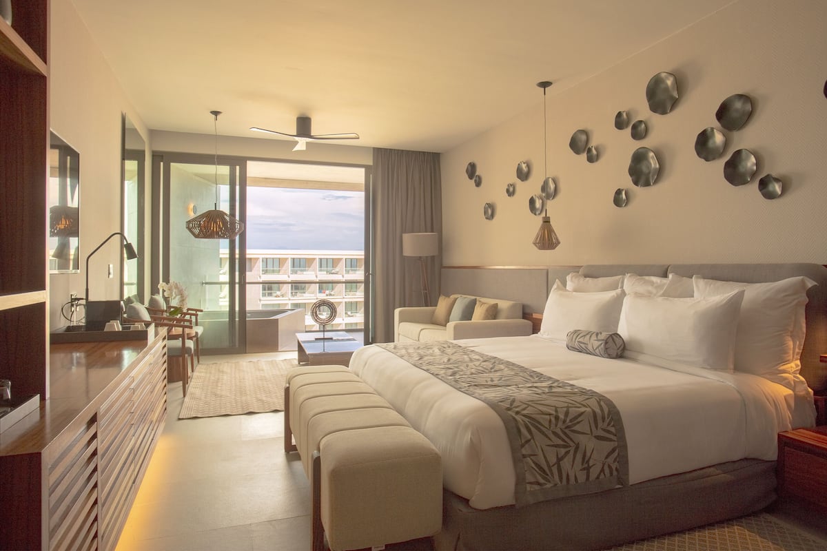 Una habitación de hotel moderna con una cama grande, una zona de estar con mesa y un balcón con vistas.