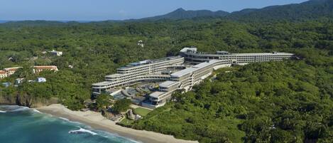 Vista aérea de un complejo turístico con playa, varias edificaciones y exuberante vegetación.