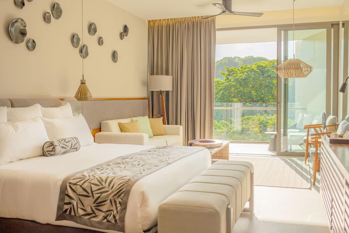 Una habitación de hotel moderna con una cama grande, un sofá, un escritorio con sillas y un balcón con vistas a zonas verdes.