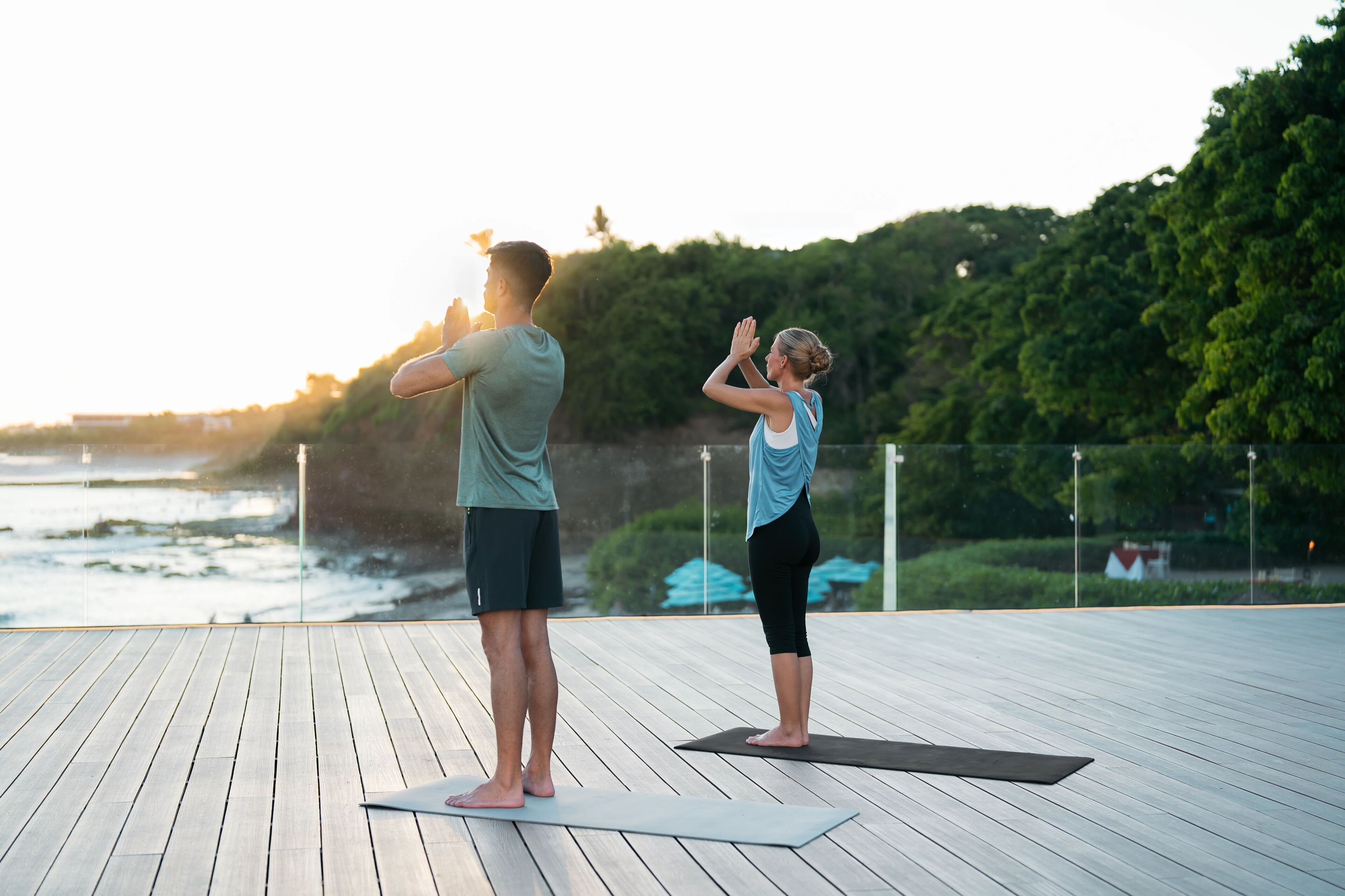 Two people practicing yoga on a wooden deck with a view of the ocean and trees in the background.