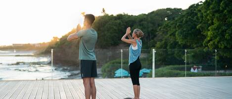 Two people practicing yoga on a wooden deck with a view of the ocean and trees in the background.