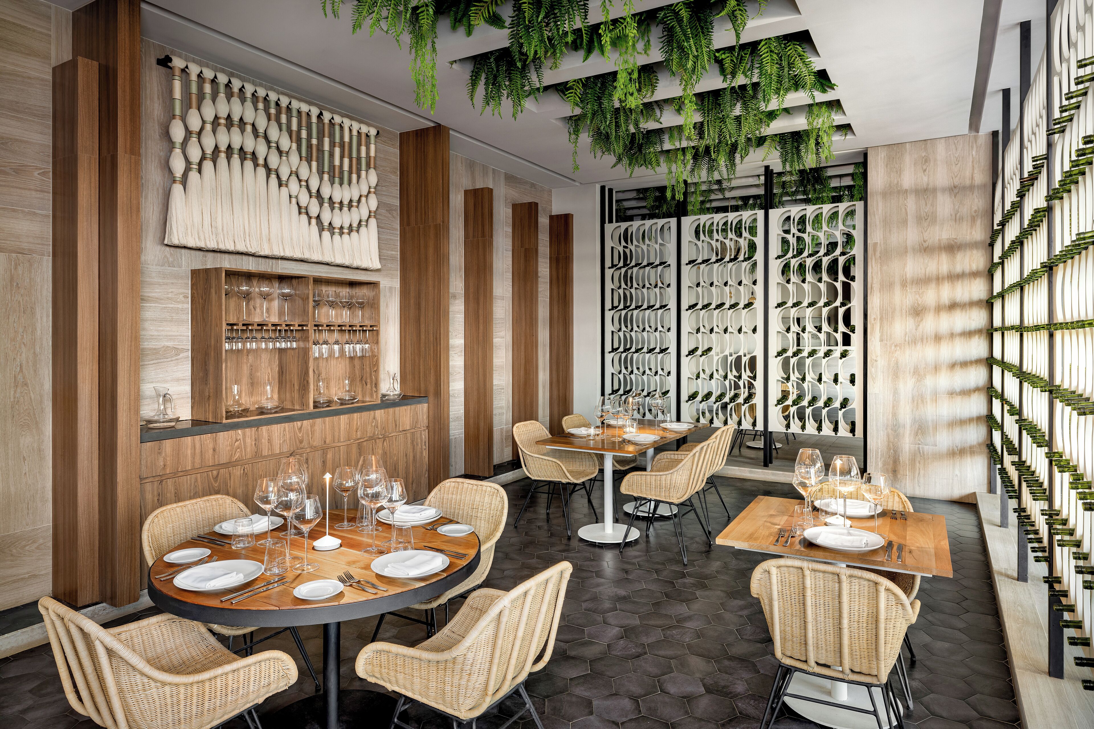 A restaurant with round tables, wicker chairs, and a wine rack on the right.