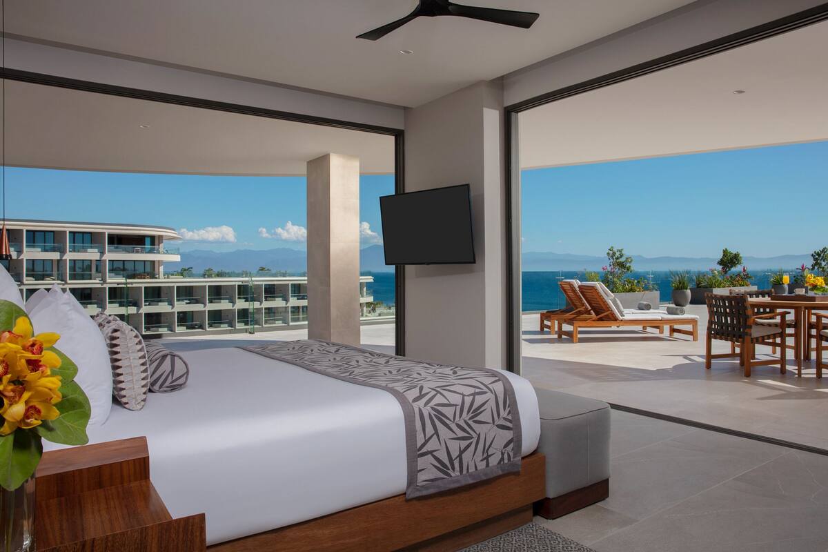 Un dormitorio moderno con una cama grande, un televisor y un balcón con vista al océano y un edificio.