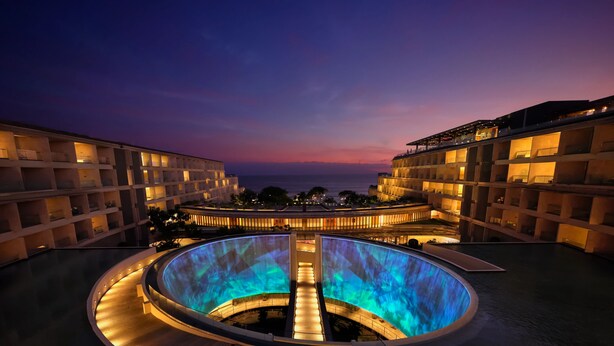 A hotel with a central pool area, multiple balconies, and a view of the ocean at dusk.
