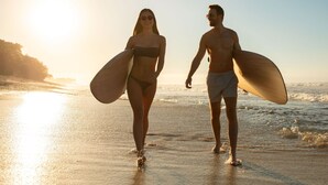 Two people walking on a beach with surfboards, the sun is setting in the background.