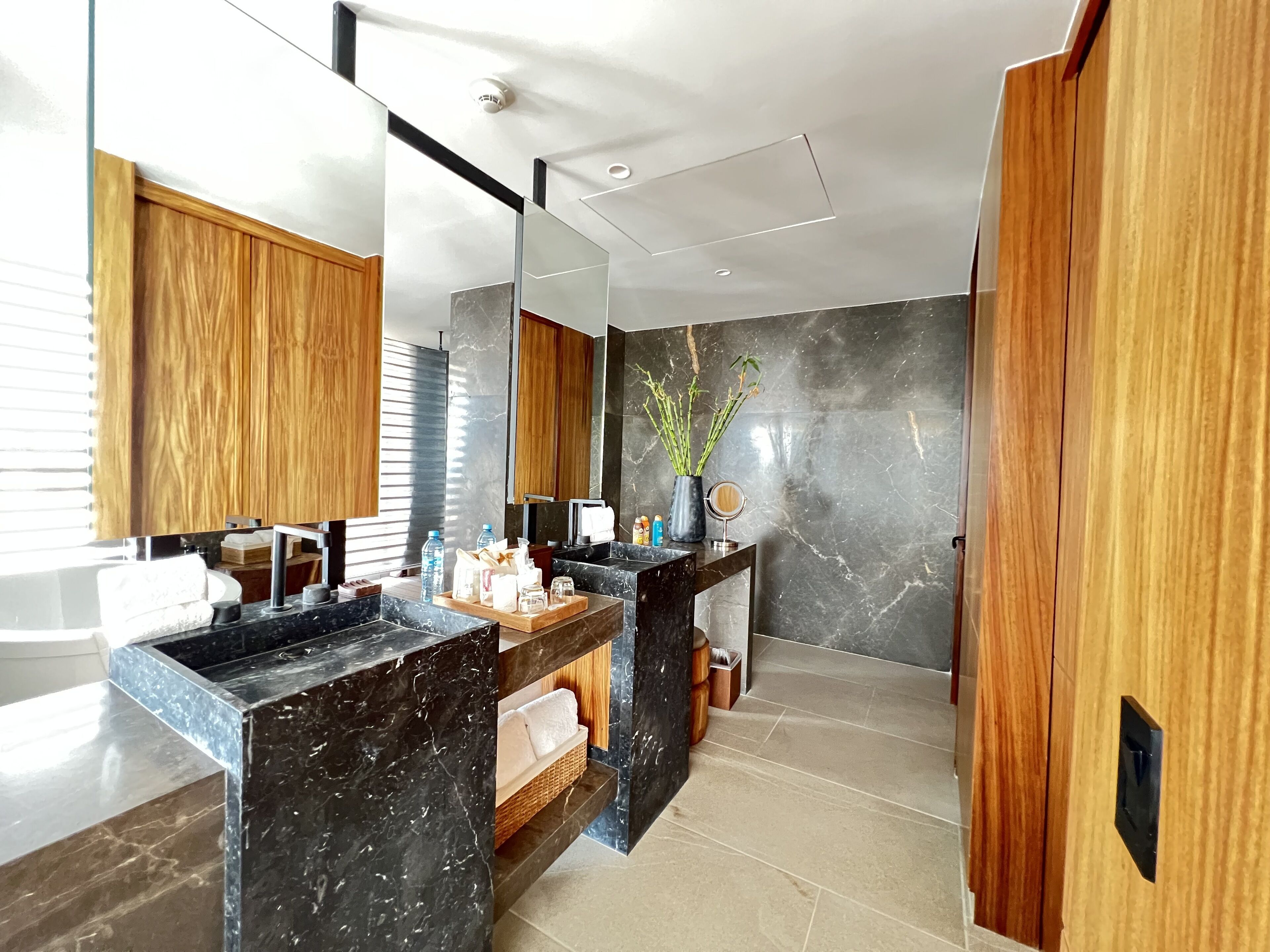 Un baño moderno con encimeras de mármol, armarios de madera, un espejo grande y un jarrón con ramas.