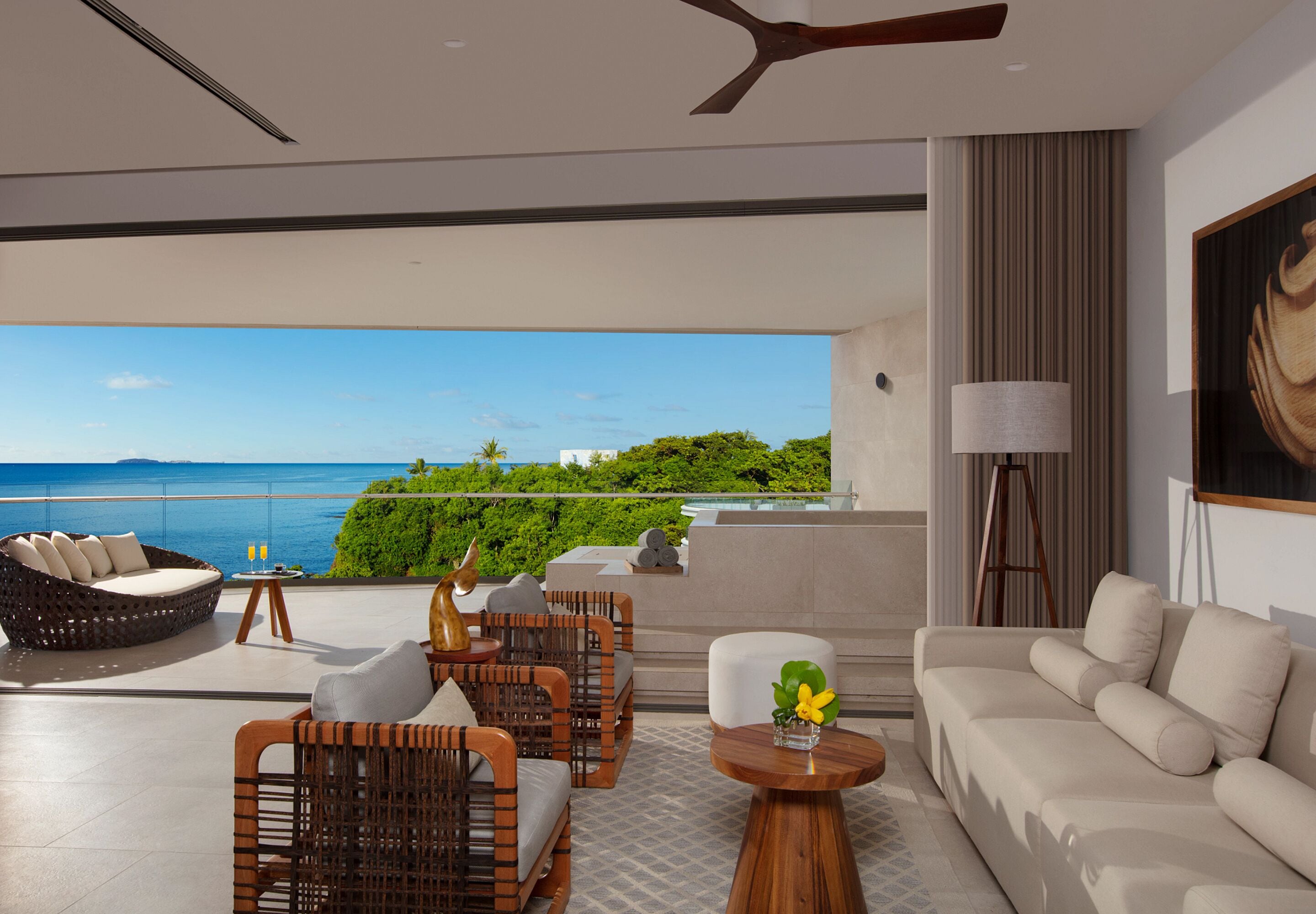 Sala de estar moderna com sofá, cadeiras e mesa, com vista panorâmica para o oceano e vegetação.
