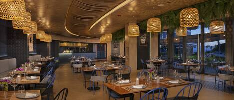 A restaurant with multiple dining tables, wicker chairs, and hanging pendant lights.