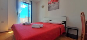 Une chambre avec un couvre-lit rouge, des oreillers blancs et une tête de lit noire. Il y a une petite table noire à côté du lit et une fenêtre avec des rideaux.