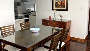 A dining area with a glass-top table, wooden chairs, a kitchen in the background, and a framed picture on the wall.