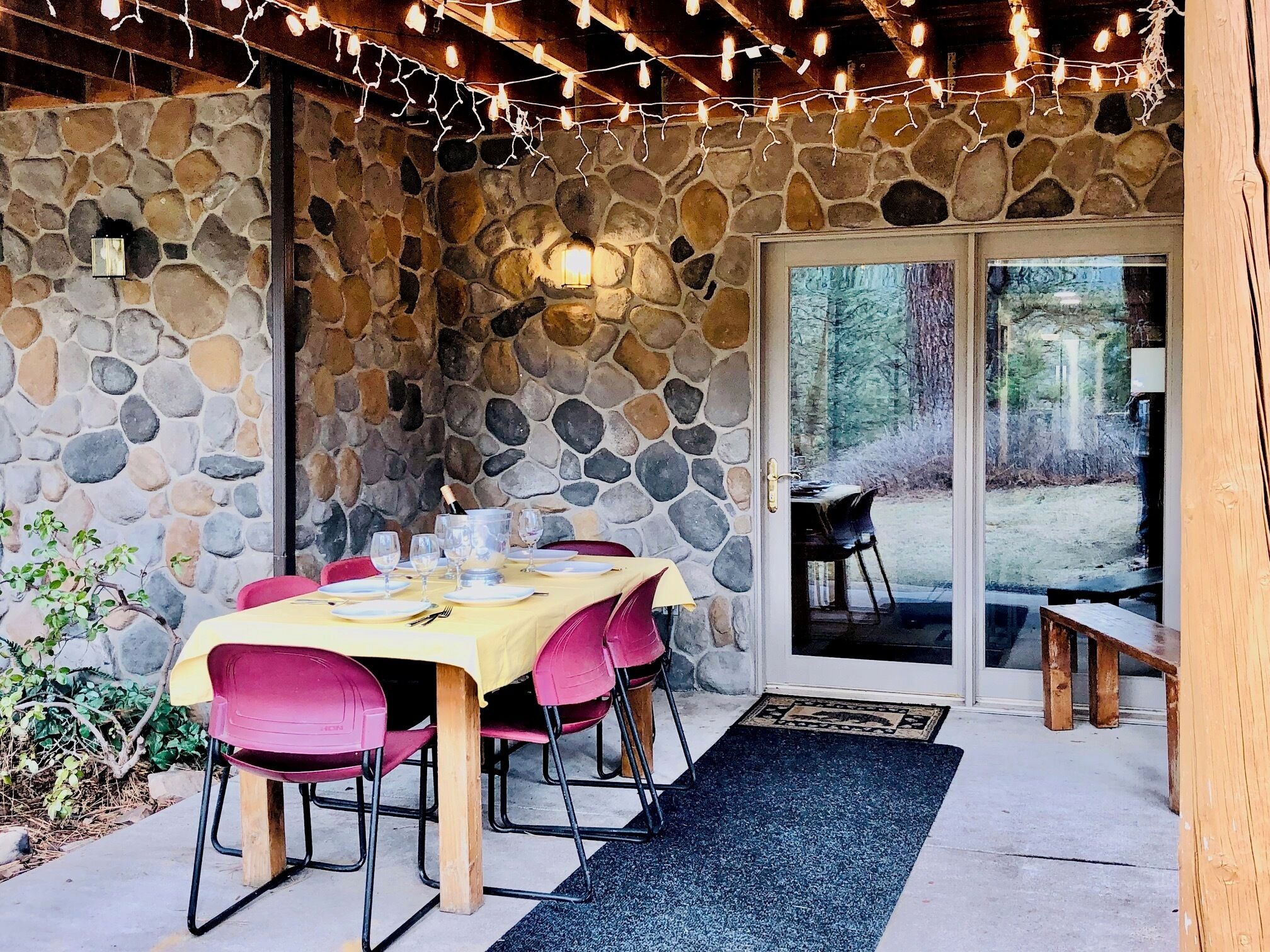 Une terrasse extérieure avec un mur en pierre, une table dressée pour le repas et des guirlandes lumineuses.