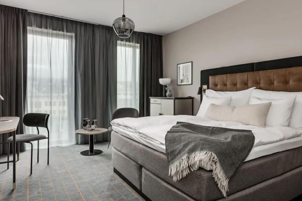 A hotel room with a large bed, a desk, a chair, a small table, and a window with curtains.