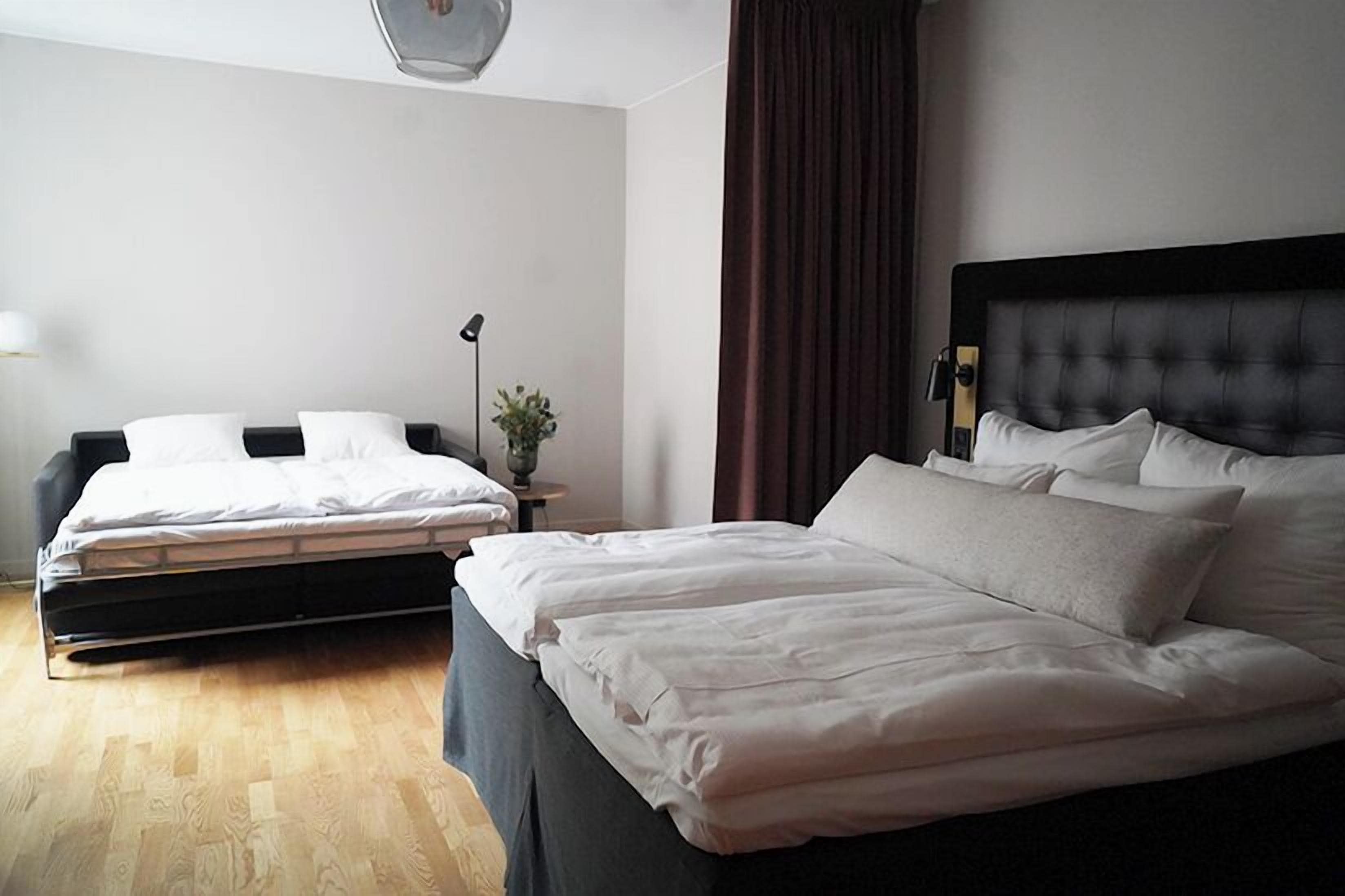 Una habitación de hotel con una cama grande, una cama más pequeña, una mesita de noche con lámpara y una planta.