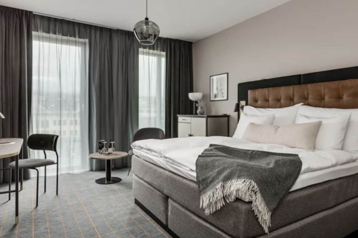 Une chambre d’hôtel avec un grand lit, un bureau, une chaise, une petite table et une fenêtre avec des rideaux.