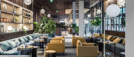 Un área de descanso moderna con sofás, sillones y mesas redondas, que combina elementos de madera y metal, y amplios ventanales que aportan luz natural.