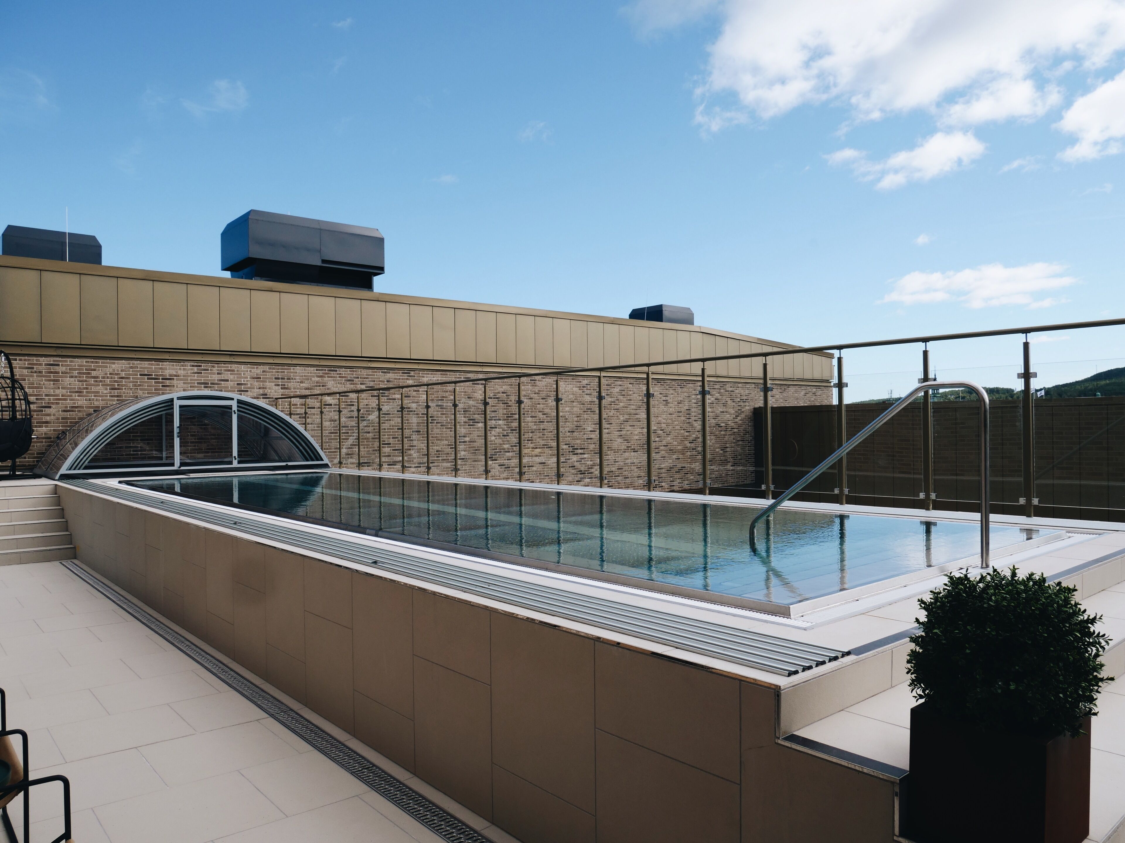 Una piscina en la azotea con cerramiento de vidrio, un muro de ladrillo y barandilla metálica.