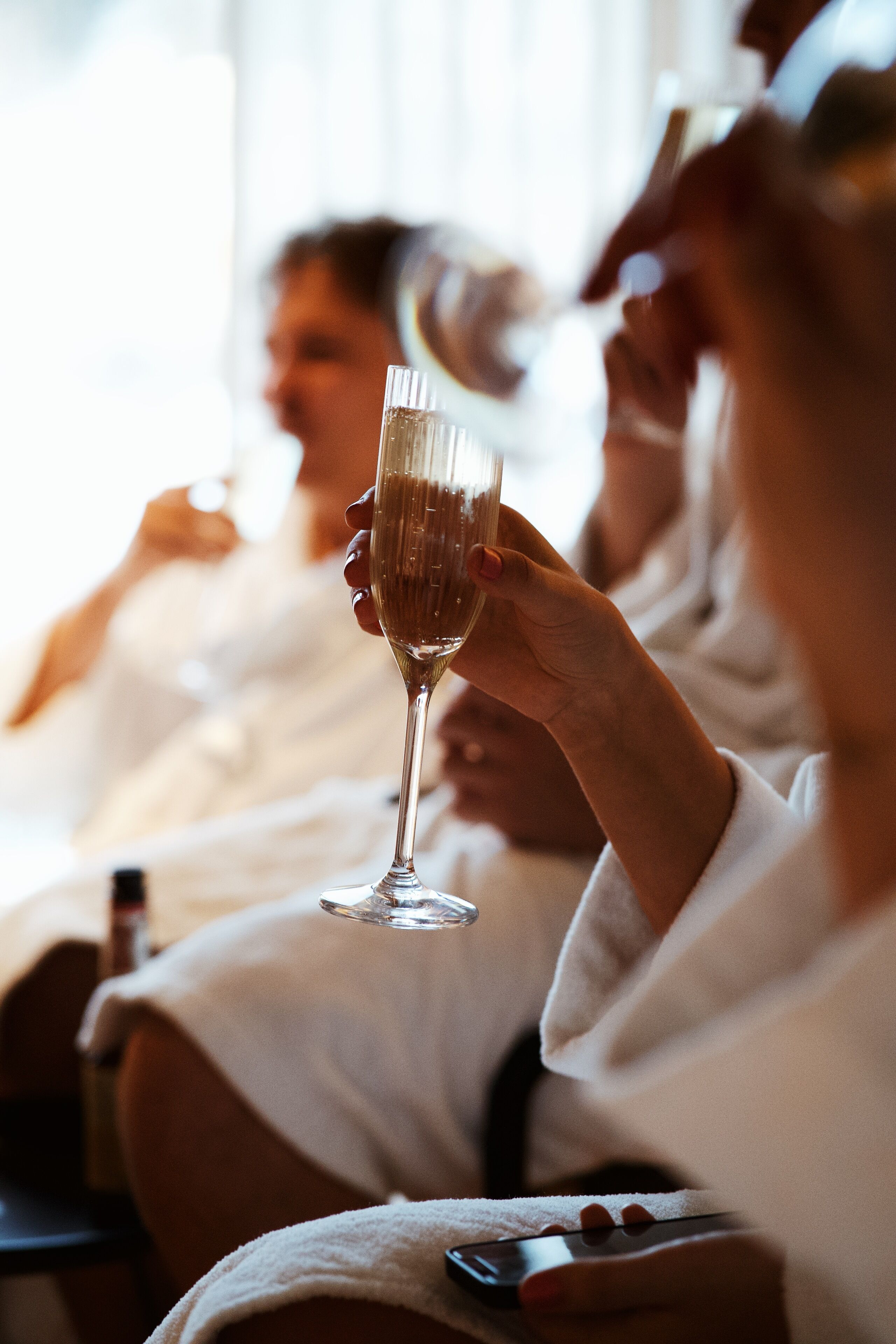 A person holding a champagne flute in a room with a window, another person in the background, and a smartphone on a table.