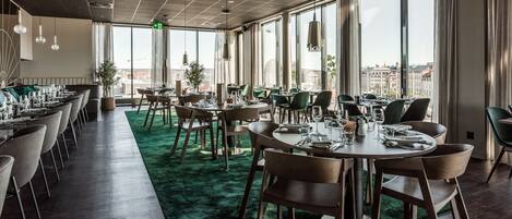 Un comedor moderno con varias mesas y sillas, ventanas amplias con vistas a la ciudad y una alfombra verde.