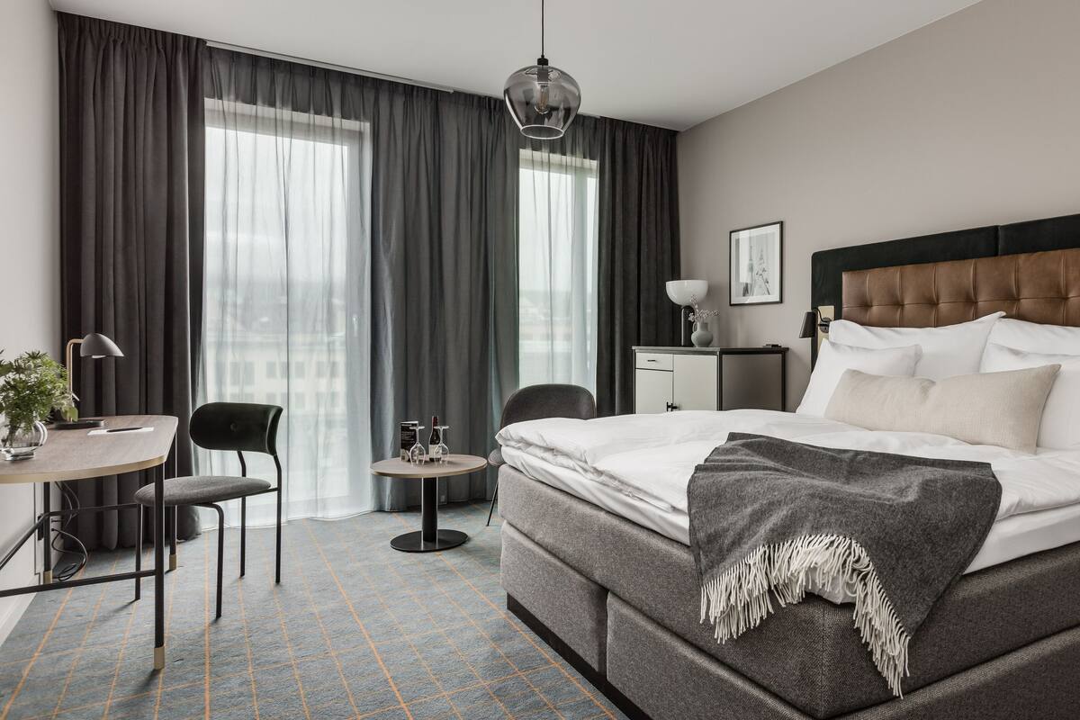 A modern hotel room with a large bed, a desk with a chair, a small table with a lamp, and a large window with curtains.