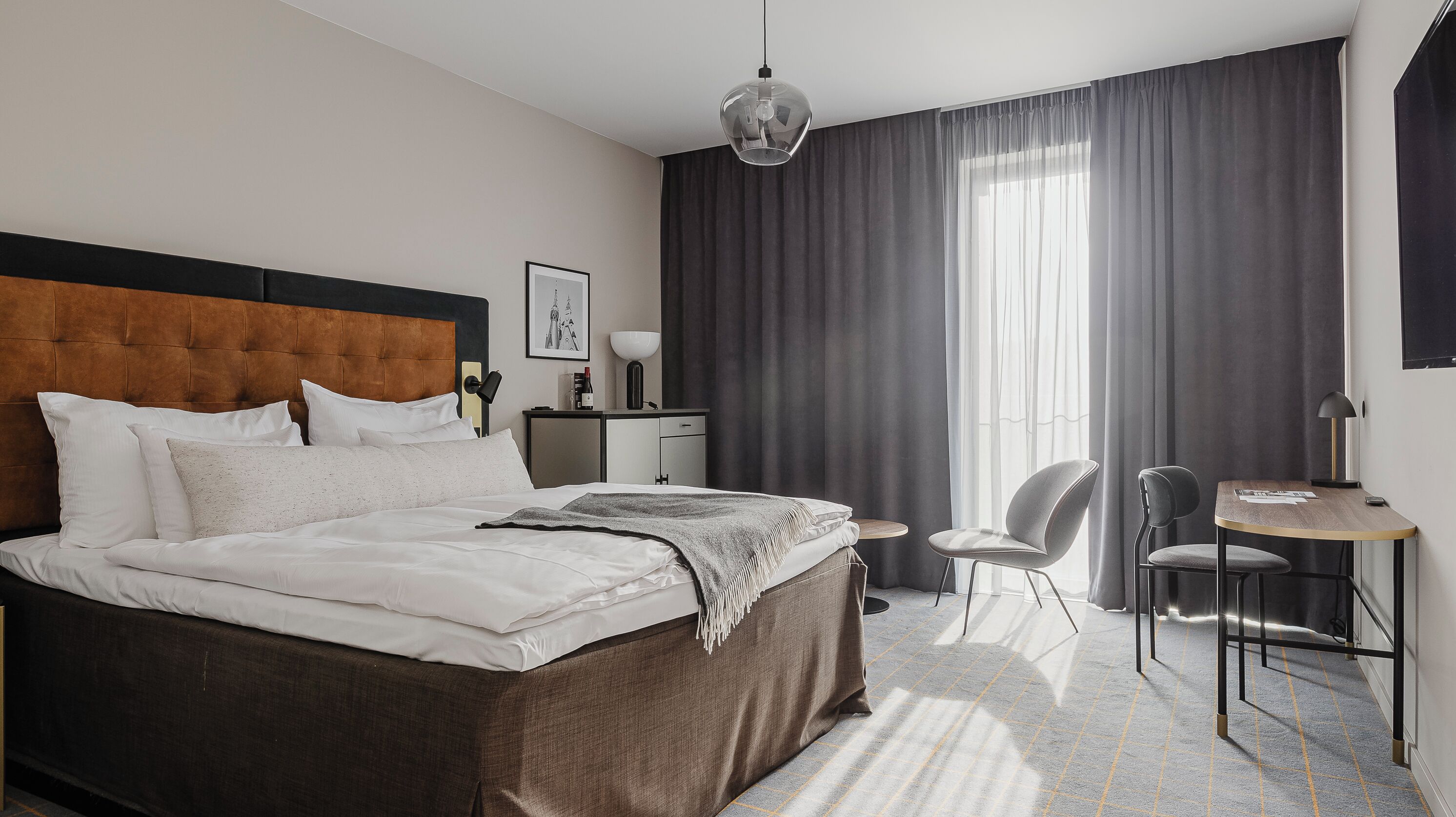 Quarto de hotel moderno com uma cama grande, uma escrivaninha, uma cadeira e vista para a cidade.