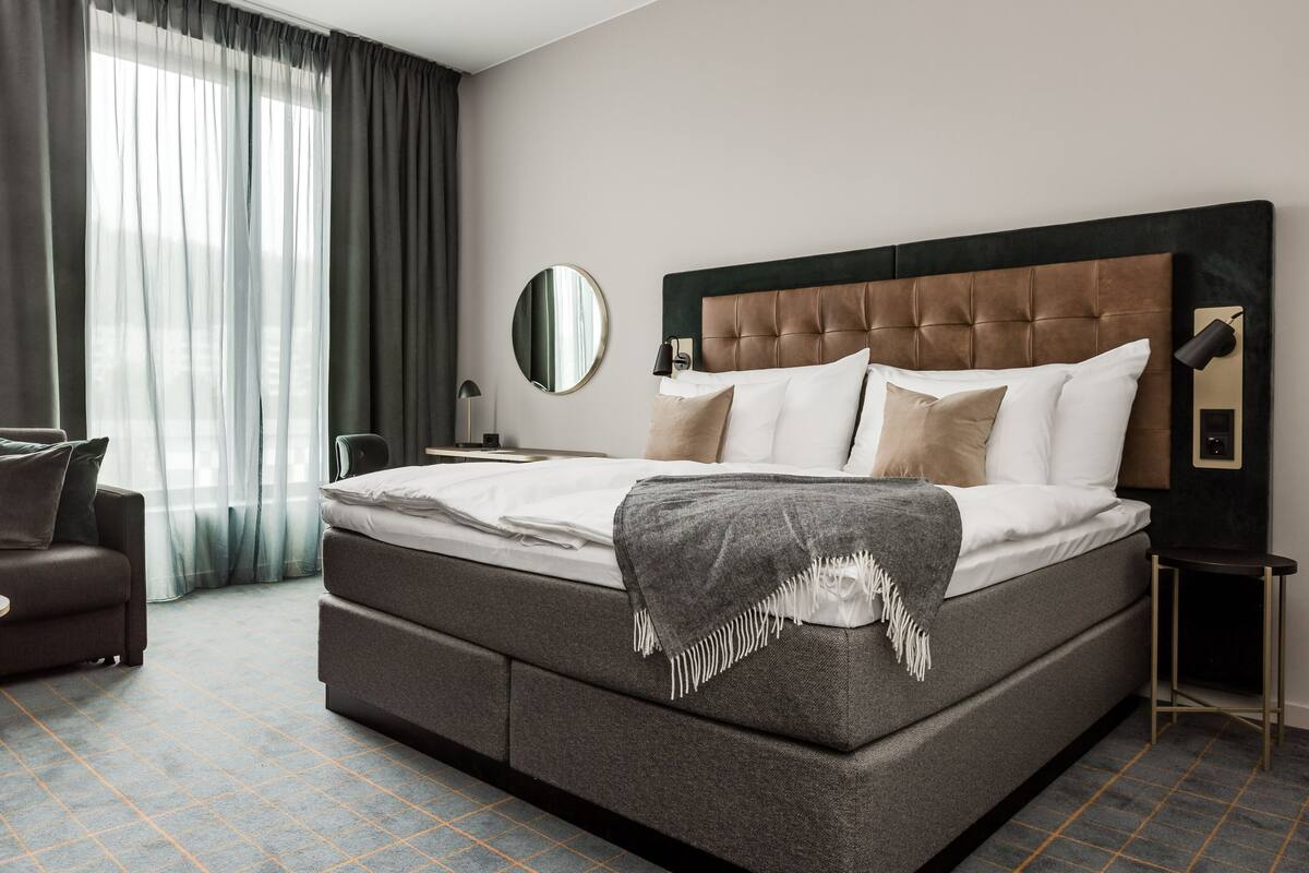 A modern hotel room with a large bed, a sofa, a round table, and a mirror.