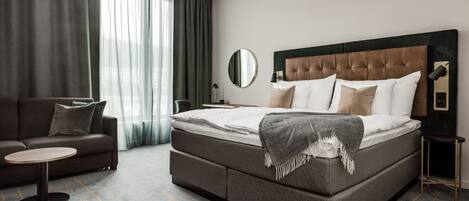 A modern hotel room with a large bed, a sofa, a round table, and a mirror.