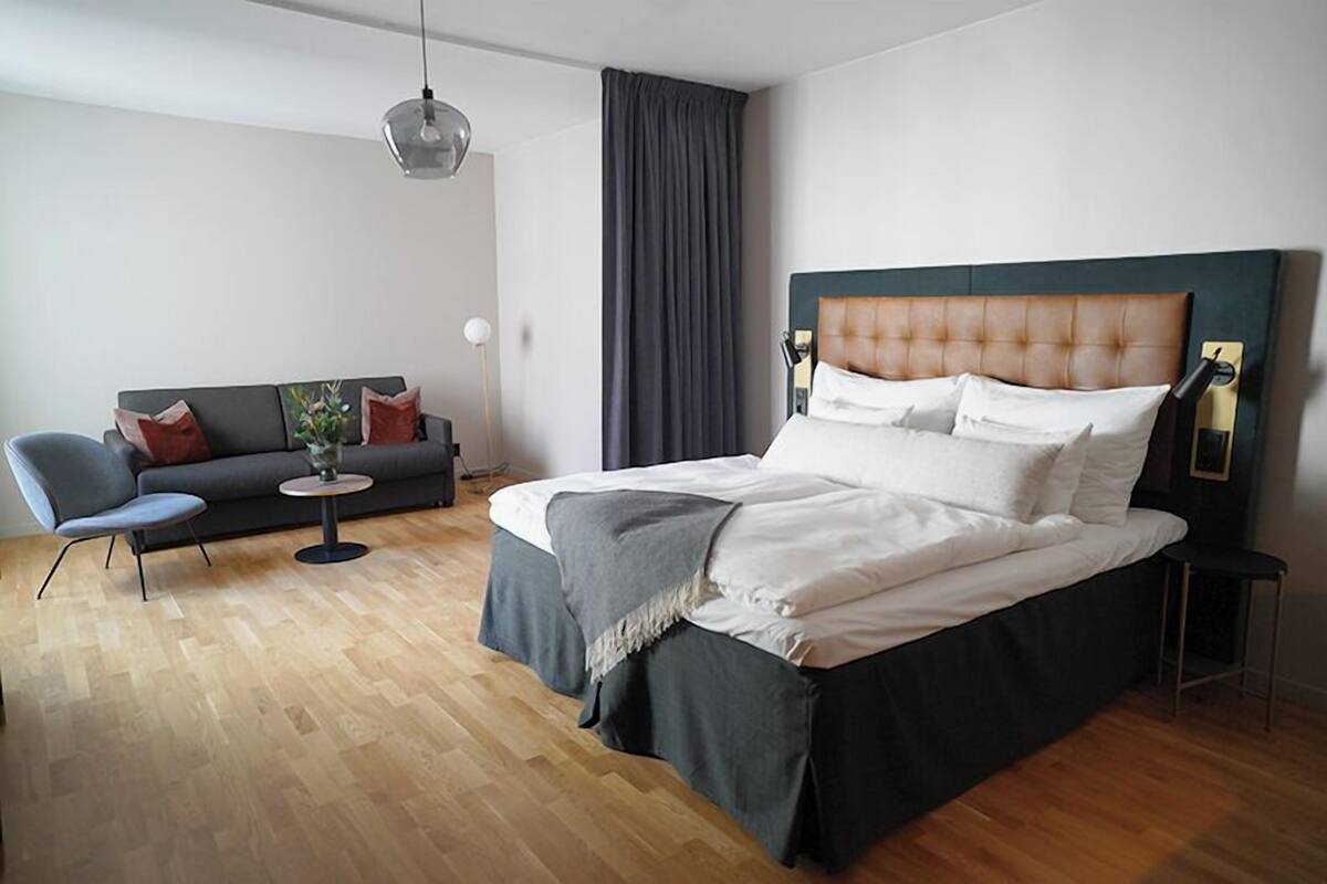 A modern hotel room with a large bed, a sofa, a small table, and a hanging light fixture.